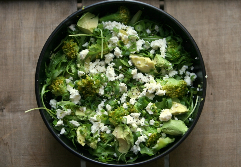 Bovenaanzicht: een zwart bord op houten tafel met daarin broccoli met avocado en feta