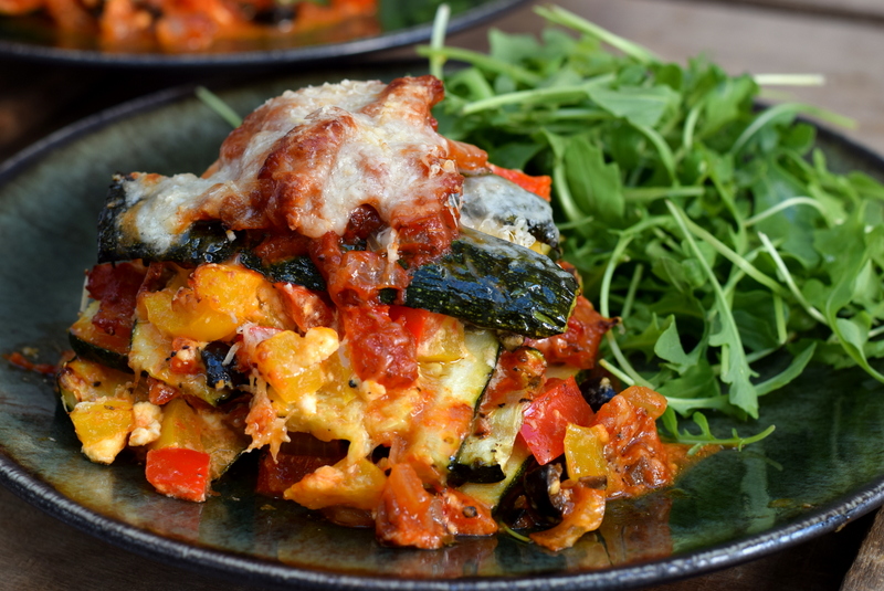 Zijaanzicht: donker bord met courgettelasagne met paprika en feta met rucola