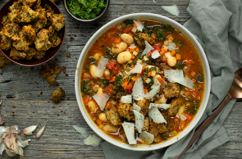 Bovenaanzicht houten tafel met rechts een kom minestrone met pestokrokjes en links extra brokjes en pesto