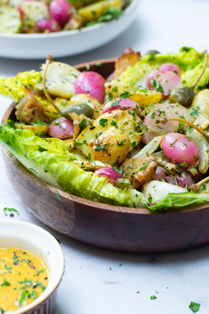 Een houten kom met felgroene Romeinse slabladeren, roze radijsjes, geroosterde krielaardappeltjes en verse kruiden. Daarnaast een potje met geel-oranje currymayonaise.