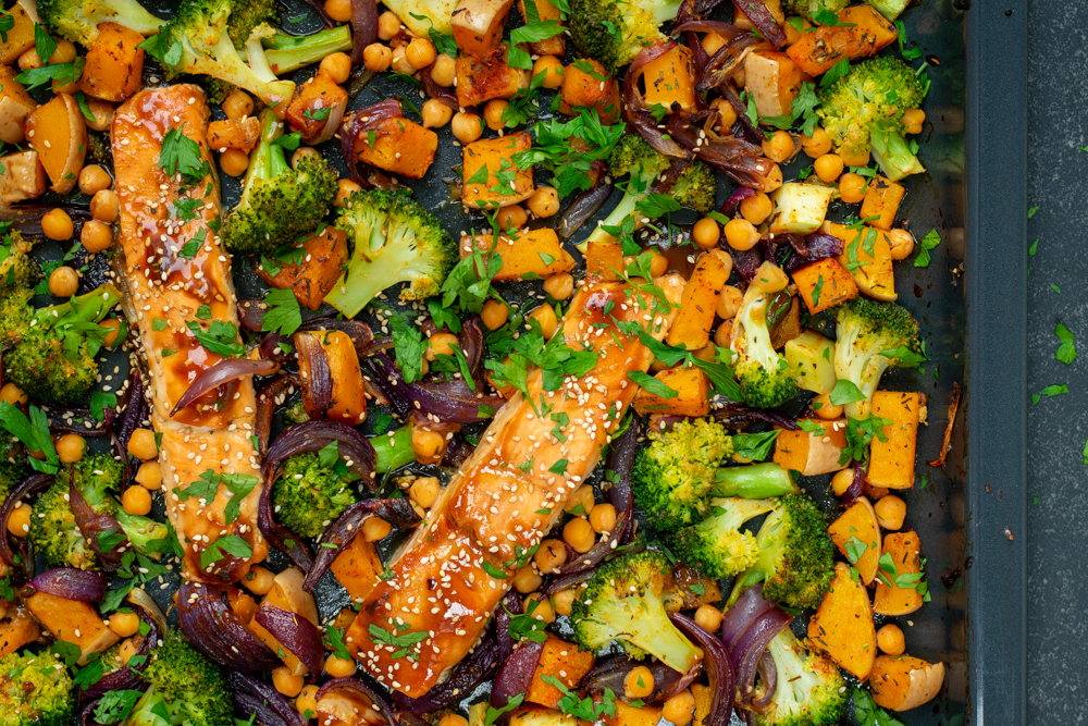 Bovenaanzicht van een bakplaat met zalm en groenten. De zalm is geglaceerd en de groenten zijn onder meer broccoli, blokjes pompoen en rode ui.