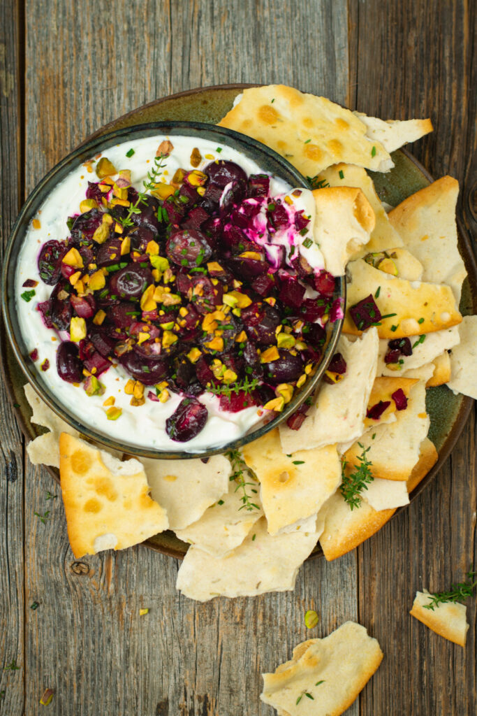 Bovenaanzicht van een dip van geitenkaas met geroosterde rode biet en druiven. Rondom de dip liggen onregelmatige, goud gebakken crackers.