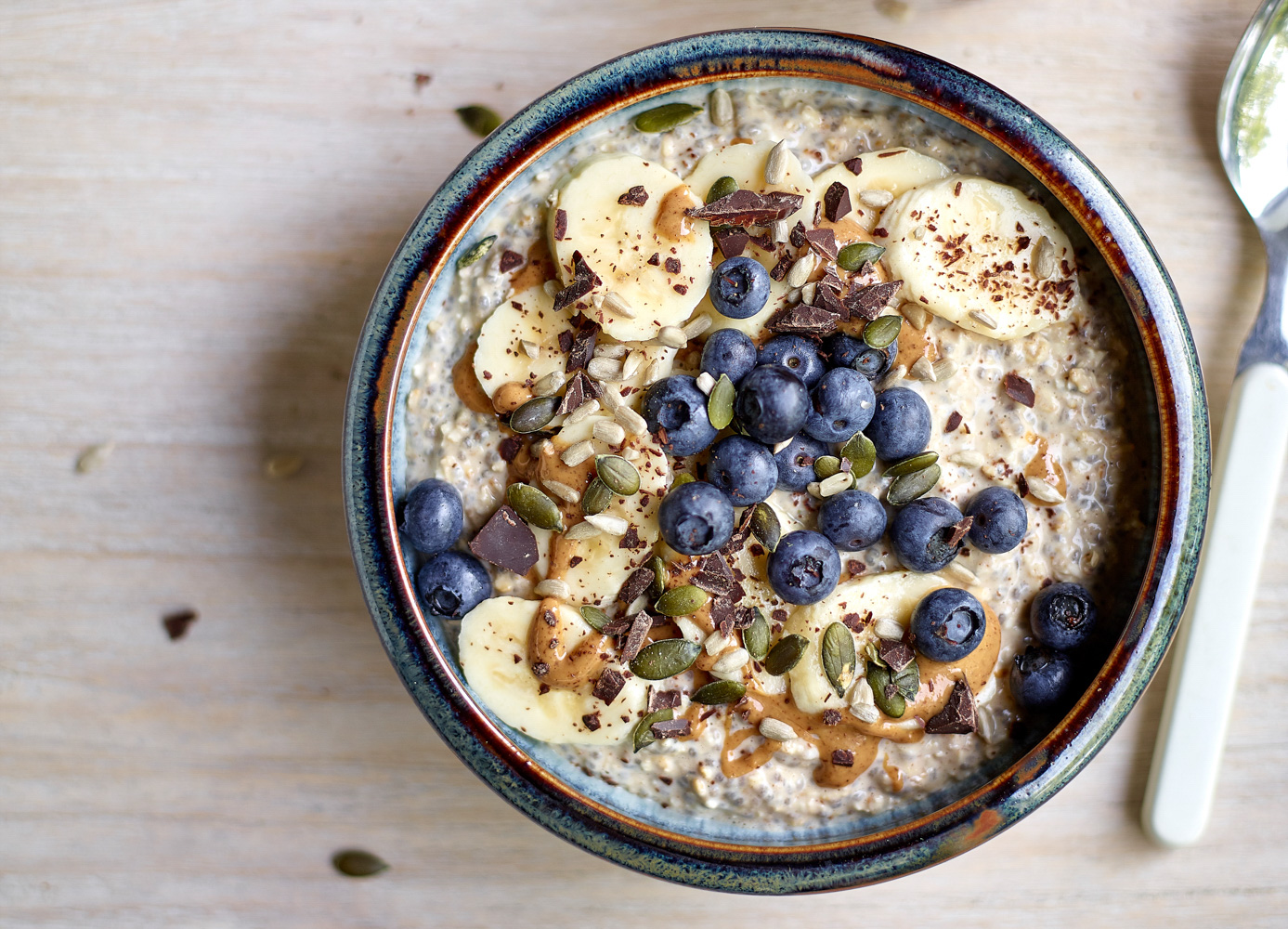 Bovenaanzicht van een kom met overnight oats, afgewerkt met schijfjes banaan, pindakaas, blauwe bessen, pompoen- en zonnebloempitten en stukjes gehakte donkere chocolade.