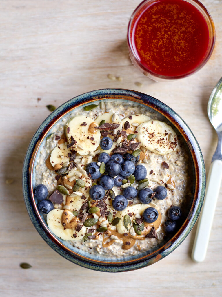 Bovenaanzicht van een kom met overnight oats, afgewerkt met schijfjes banaan, pindakaas, blauwe bessen, pompoen- en zonnebloempitten en stukjes gehakte donkere chocolade. Rechts bovenaan staat een glas met een rodebietensapje.