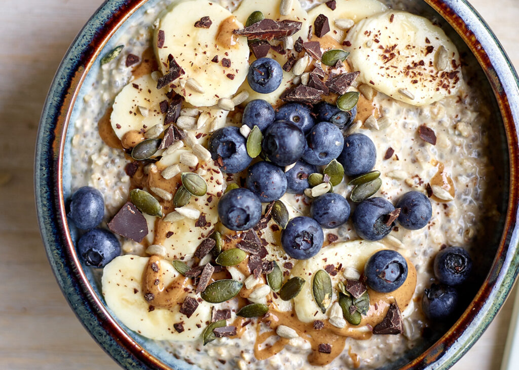 Bovenaanzicht van een kom met overnight oats, afgewerkt met schijfjes banaan, pindakaas, blauwe bessen, pompoen- en zonnebloempitten en stukjes gehakte donkere chocolade.
