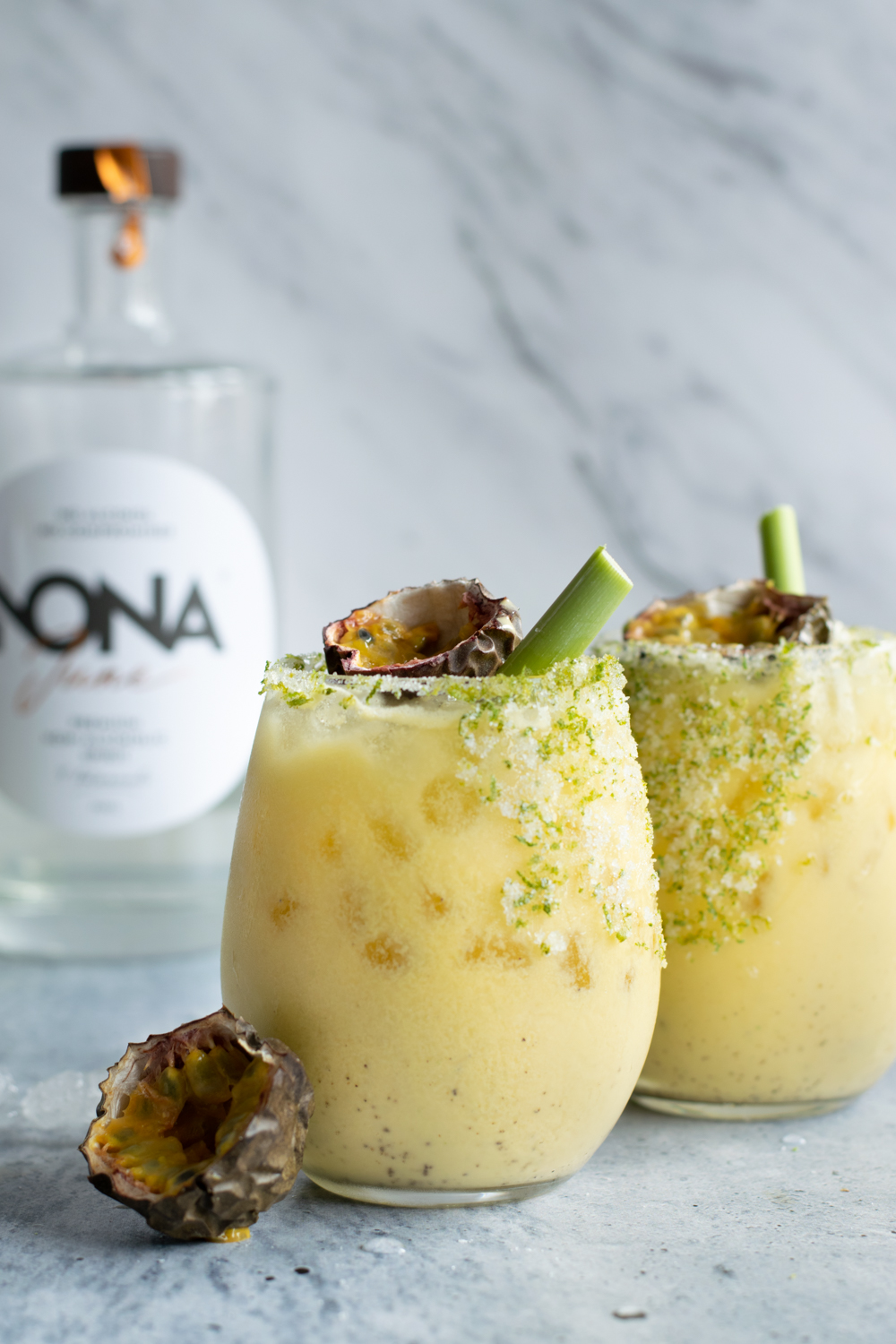 Twee bolle glazen, gevuld met een lichtgele kokos-passievruchtmocktail. De glazen zijn afgewerkt met een randje van suiker, zout en limoenzeste. De mocktail is afgewerkt met een citroengrasstengel en een halve passievrucht.