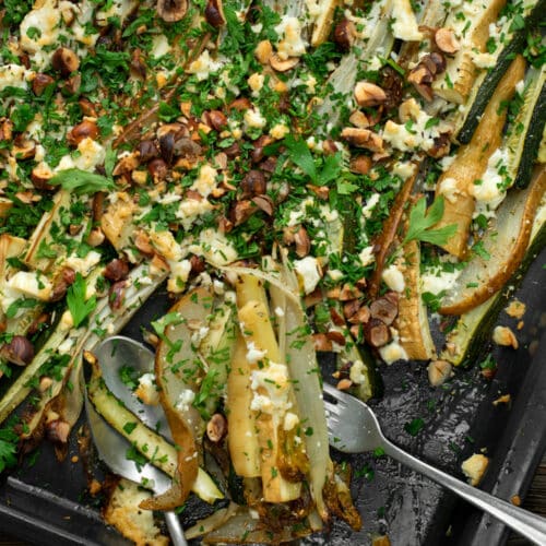 Bovenaanzicht van een bakplaat met geroosterde witloof, pastinaak en peer, afgewerkt met groene kruiden, hazelnoten en feta.