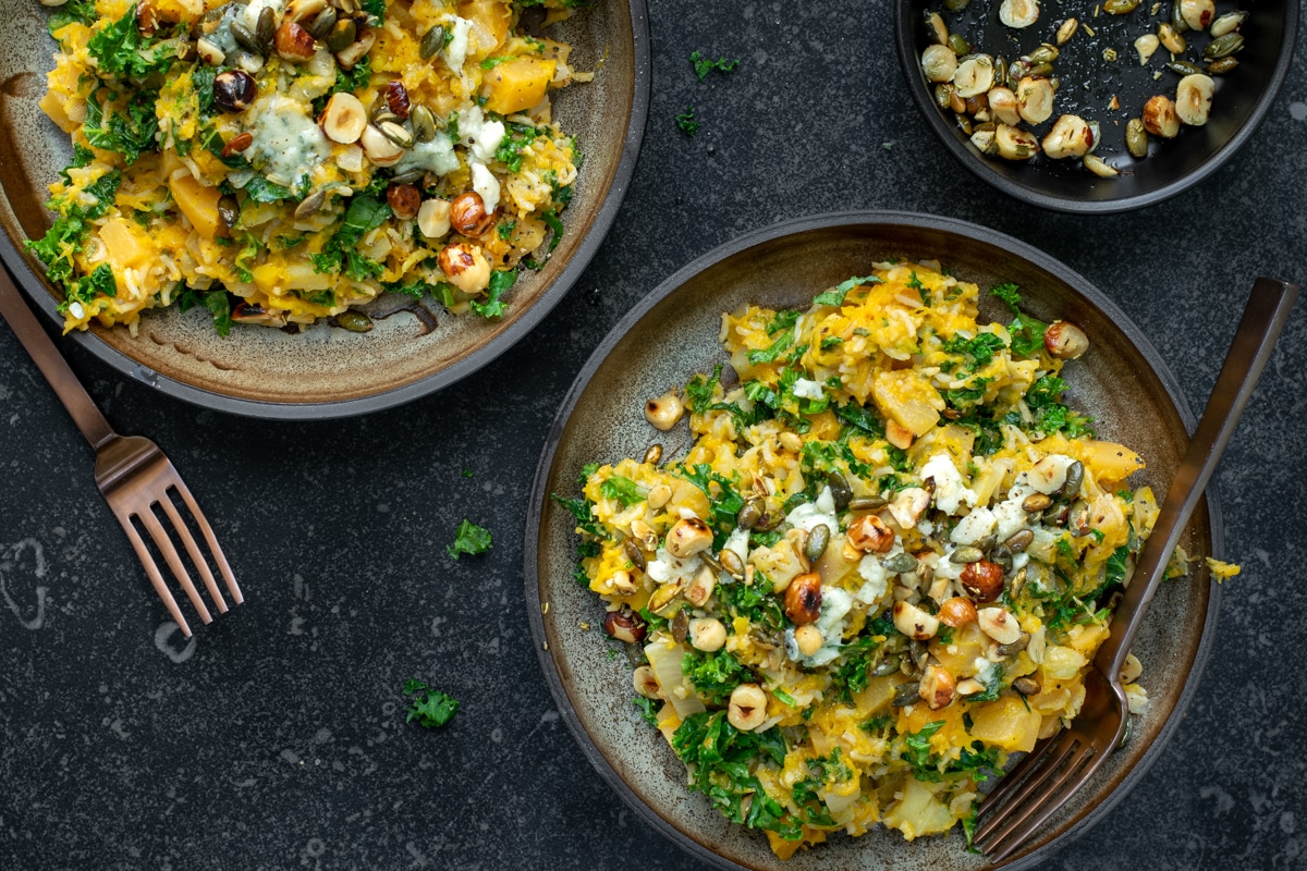 Bovenaanzicht van twee donkere borden op een donkere achtergrond. De borden zijn gevuld met een oranjekleurige pompoenrisotto, afgewerkt met blauwe kaas en noten.